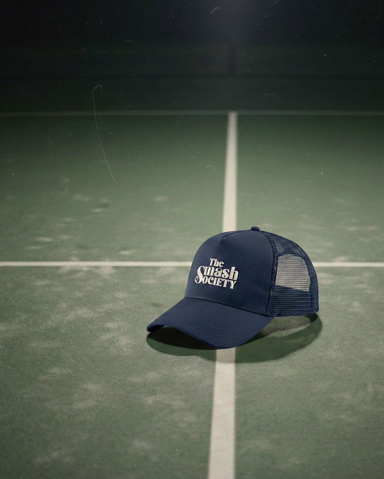 a navy blue trucker padel with the smash society logo embroidered on it the cap laying on a padel court