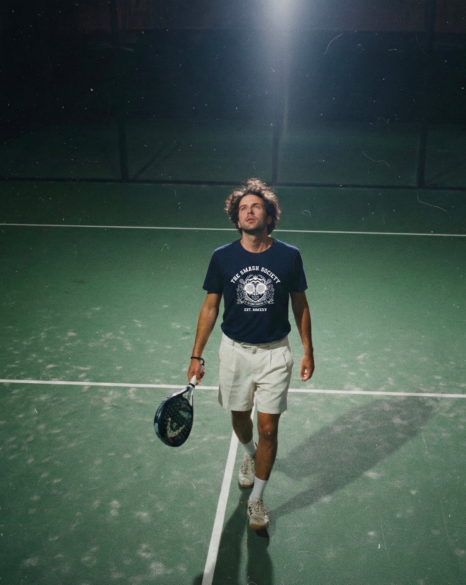 Man walking on a padel court holding a racquet at night and wearing a the smash society navy padel t-shirt