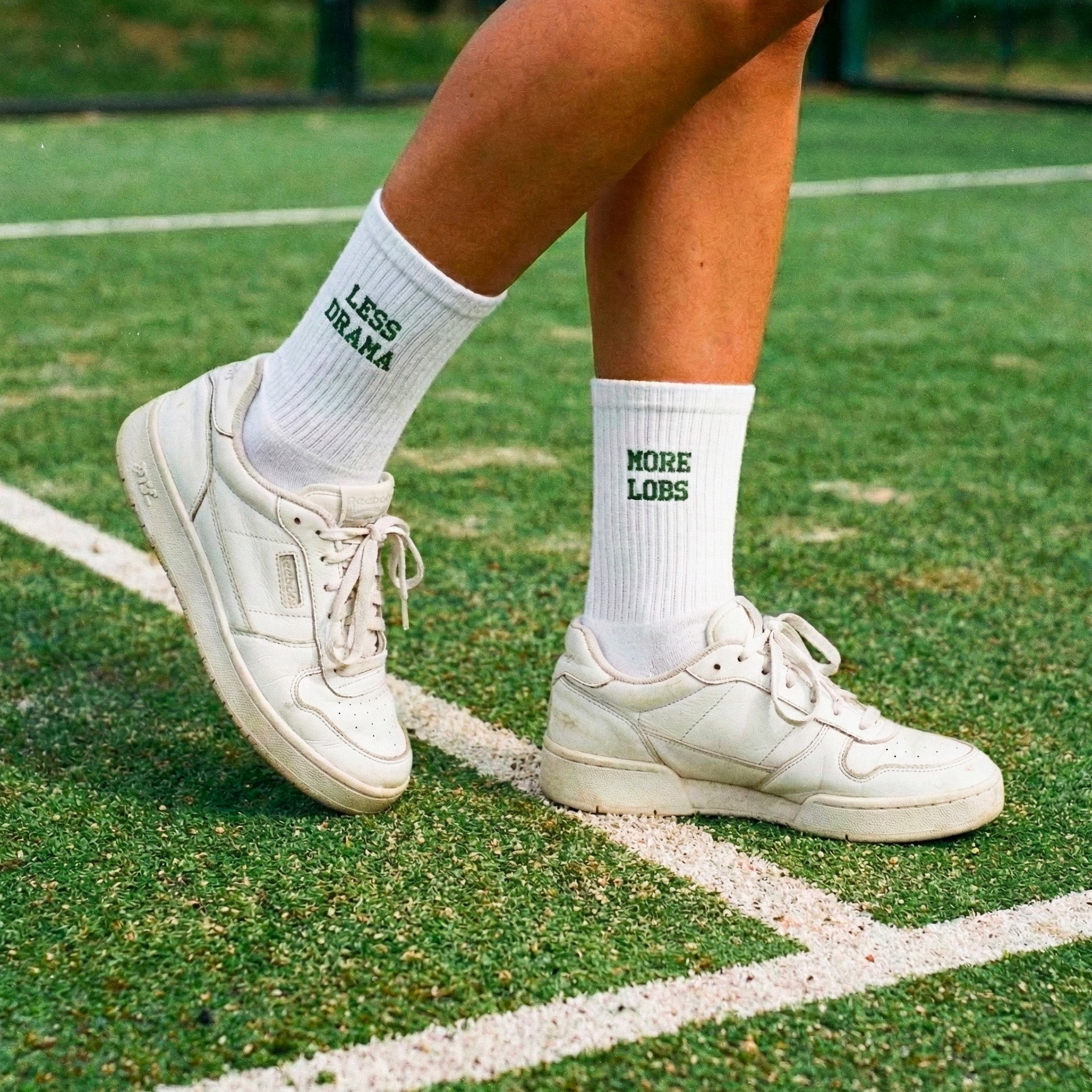 White sneakers and socks with more lobs less drama on them on a grassy padel field