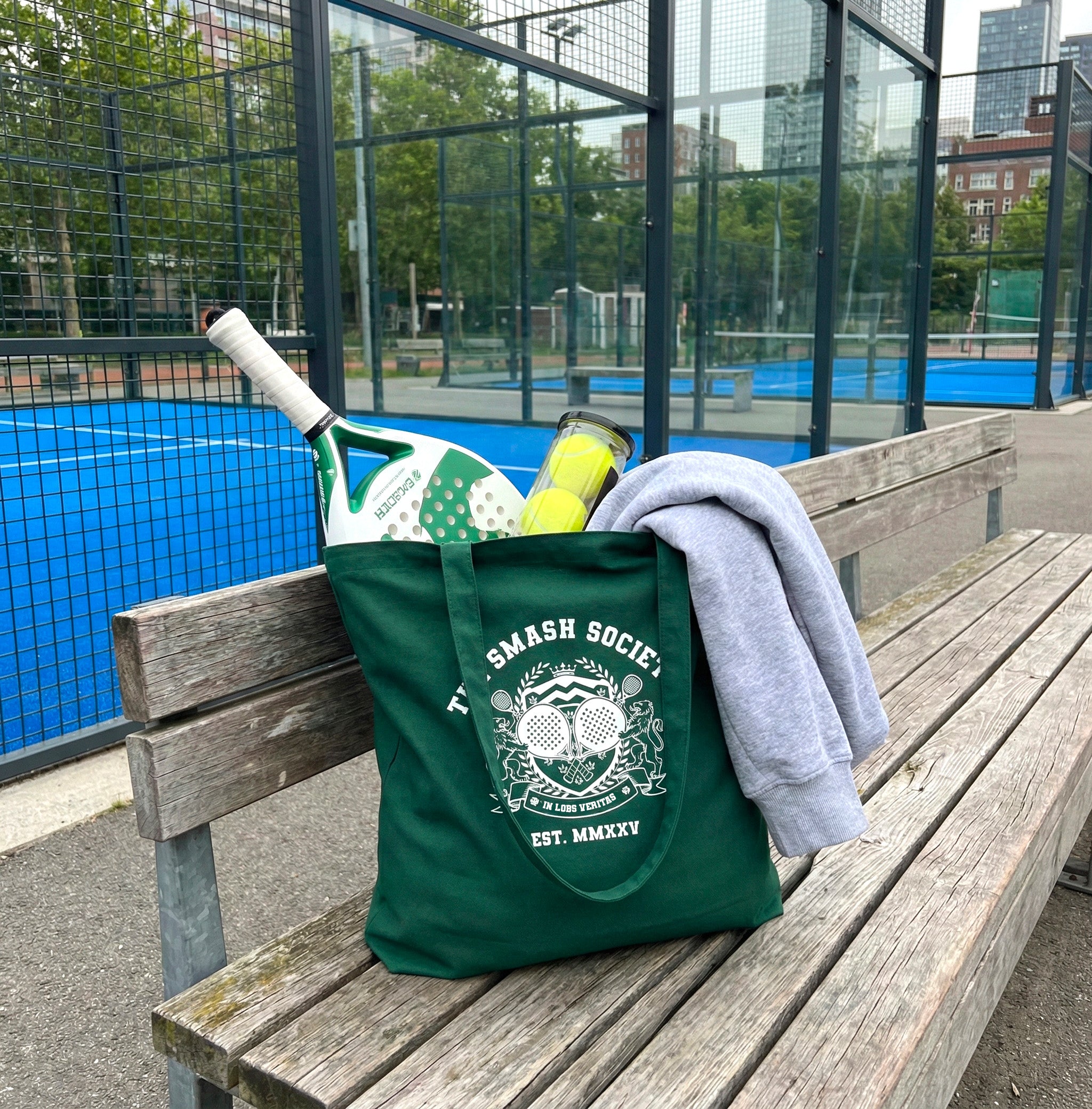 Green tote bag with 'The Smash Society' logo on a bench near a padel court