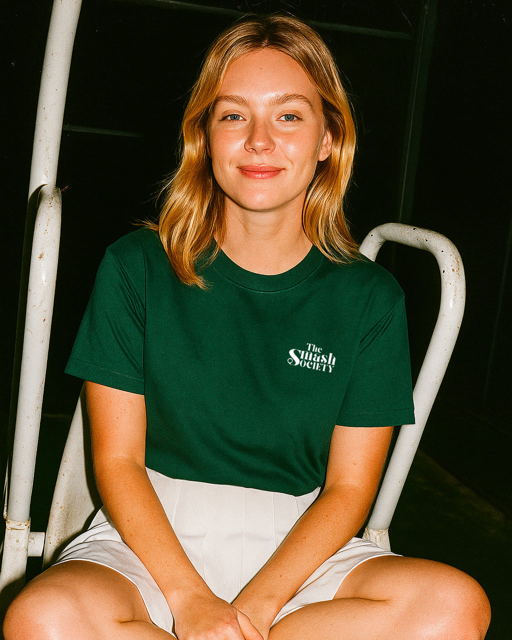 Person wearing a green t-shirt with text sitting on a white chair against a dark background