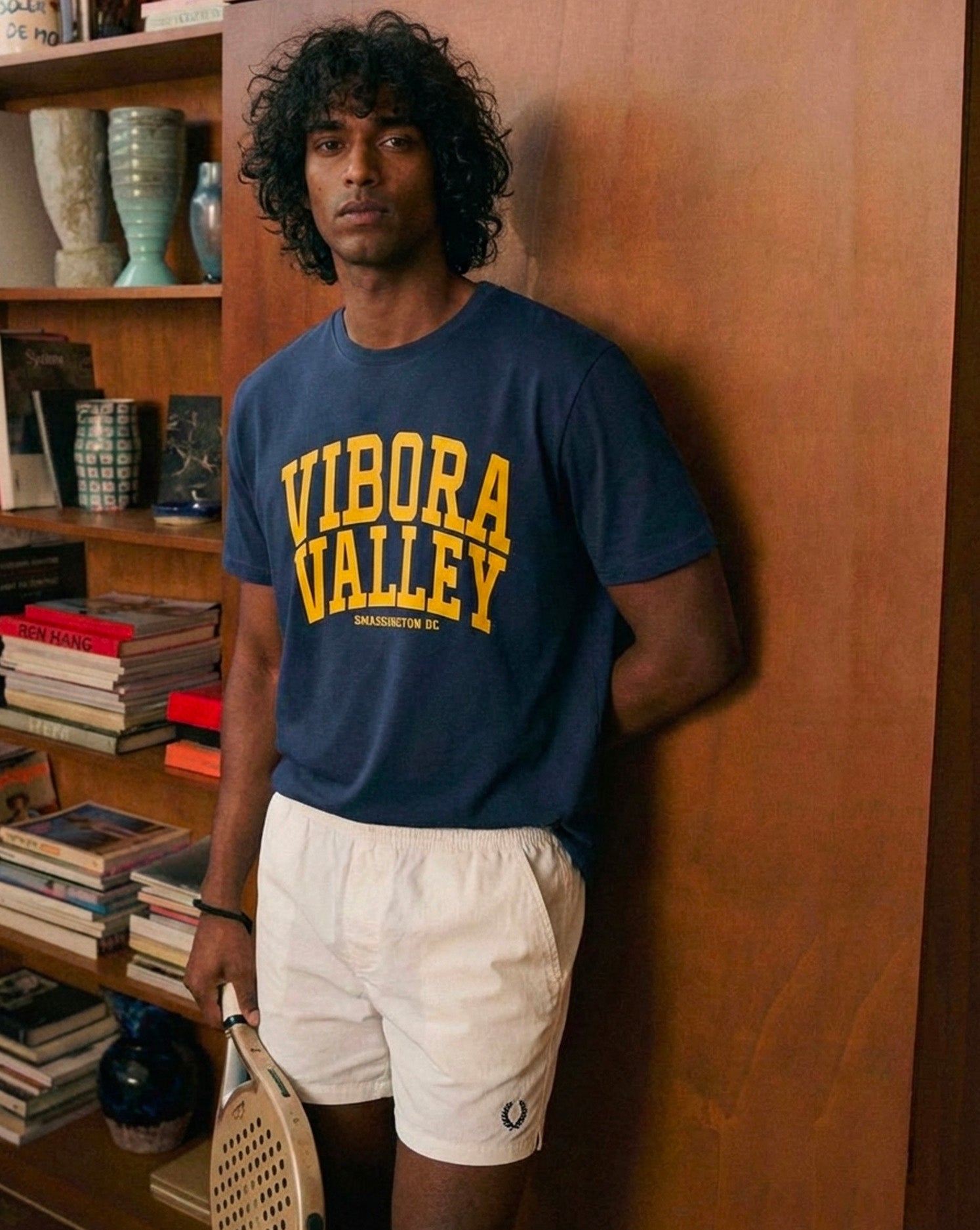 Man wearing a blue 'Vibora Valley' t-shirt and white shorts, holding a padel racket, against a wooden wall. the smash society