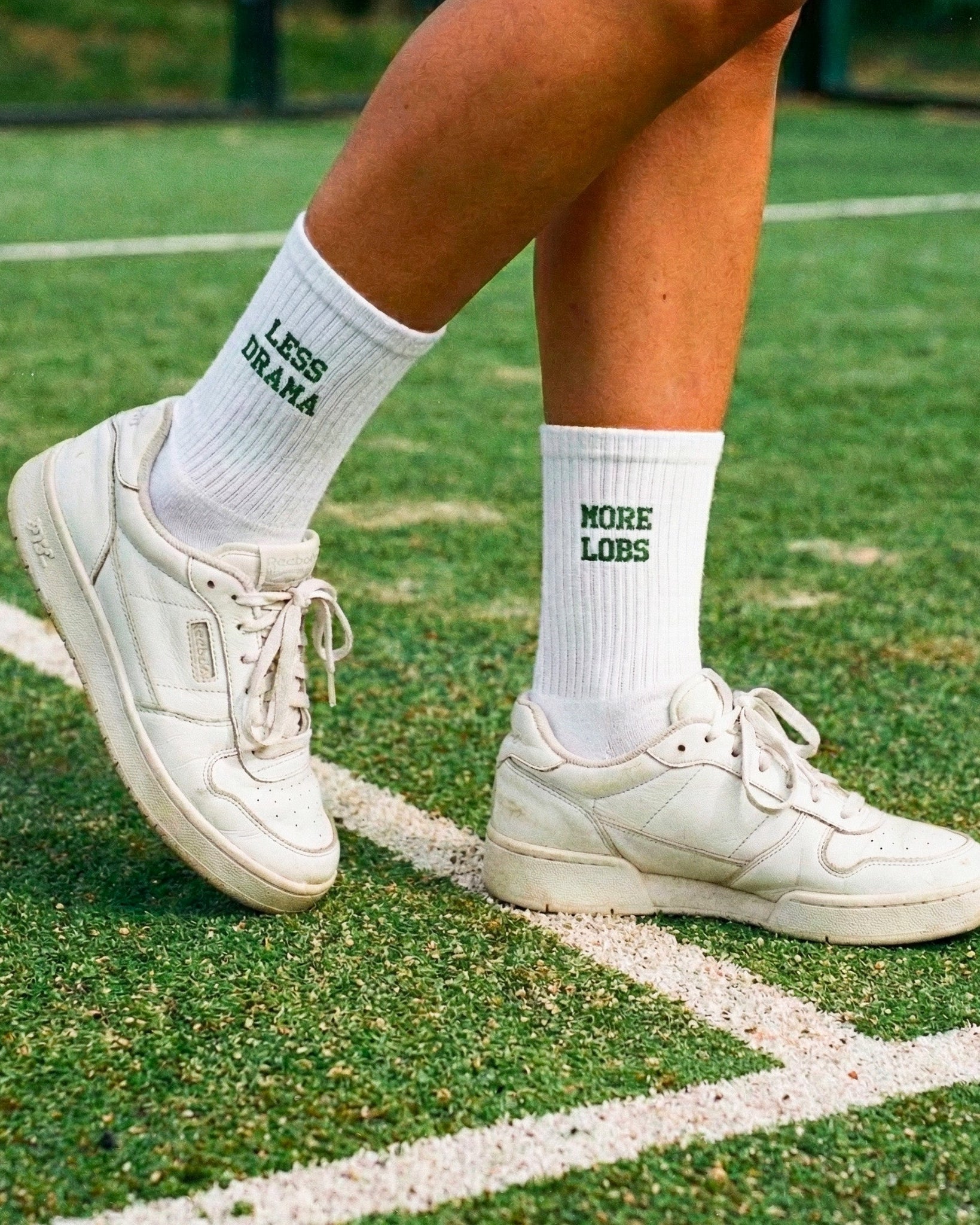 White sneakers and socks with more lobs less drama on them on a grassy padel field