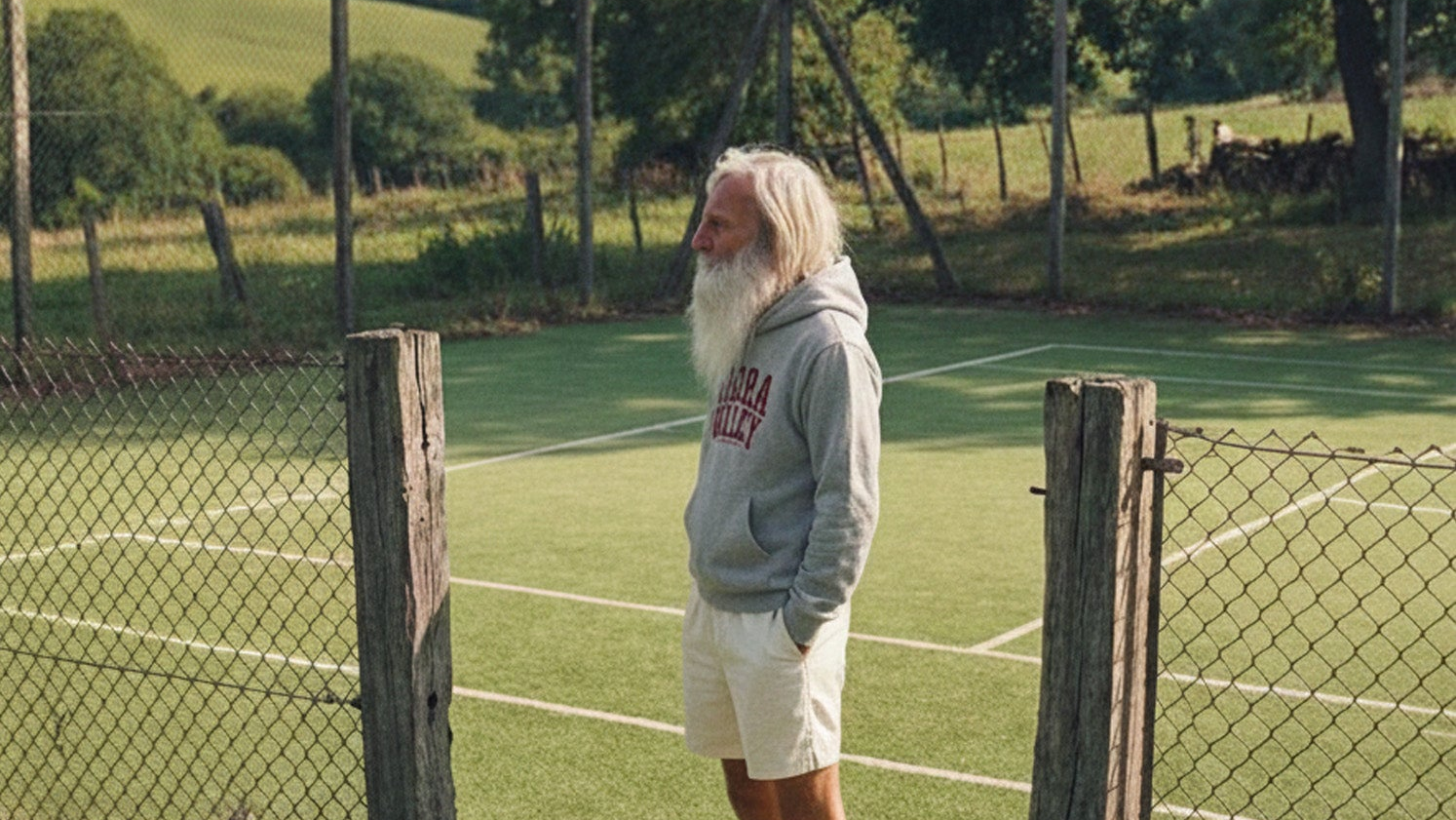 Man with a long white beard and a grey 'vibora valley' sweatshirt standing on a tennis court with a fence and trees in the background.